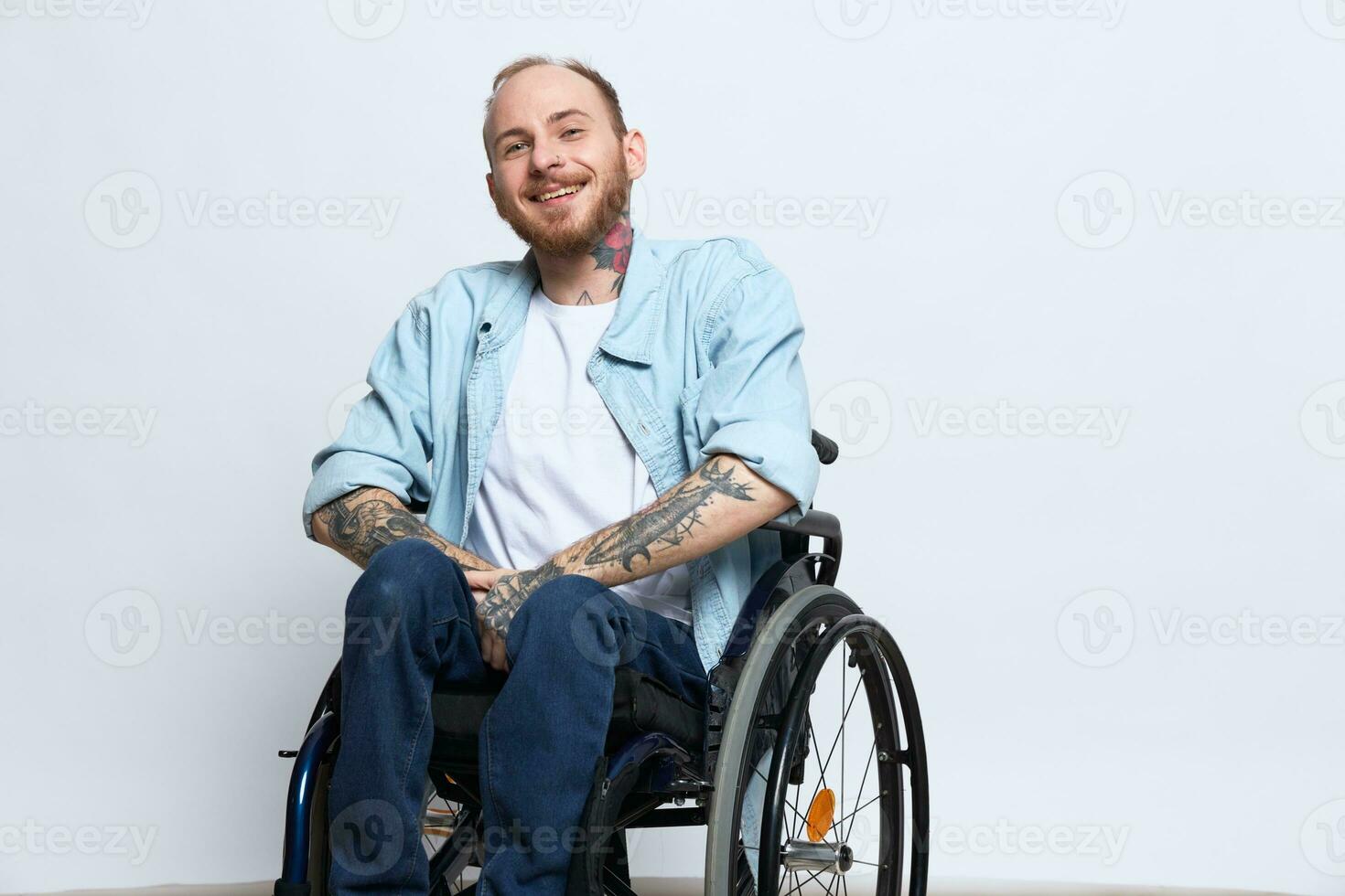 A man in a wheelchair and pointing a finger at, copy space, with tattoos on his hands sits on a gray studio background, health concept a person with disabilities, a real person photo