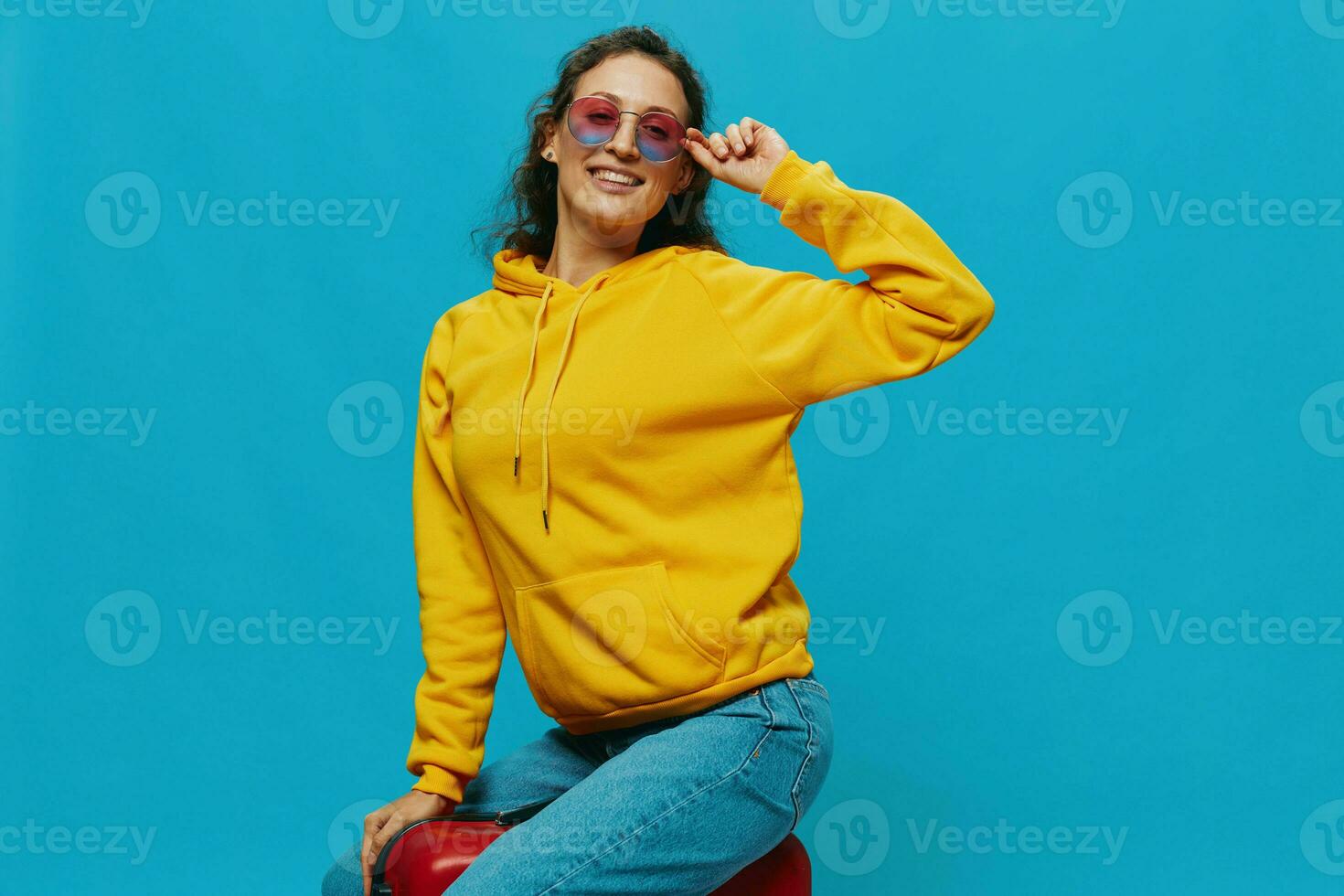 Smile woman sitting on a suitcase in a yellow hoodie, blue jeans and glasses on a blue background, packing for a trip photo