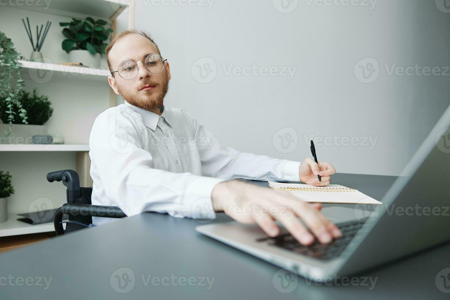 A man wheelchair businessman in the office works at a laptop, writing down ideas and a plan in a notebook, thoughtfulness, integration into society, the concept of working a person with disabilities photo