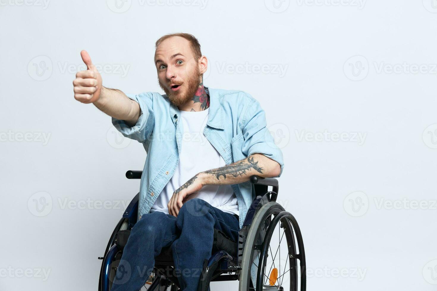 A man in a wheelchair looks at the camera shows a thumbs up, happiness, with tattoos on his hands sits on a gray studio background, health concept man with disabilities photo