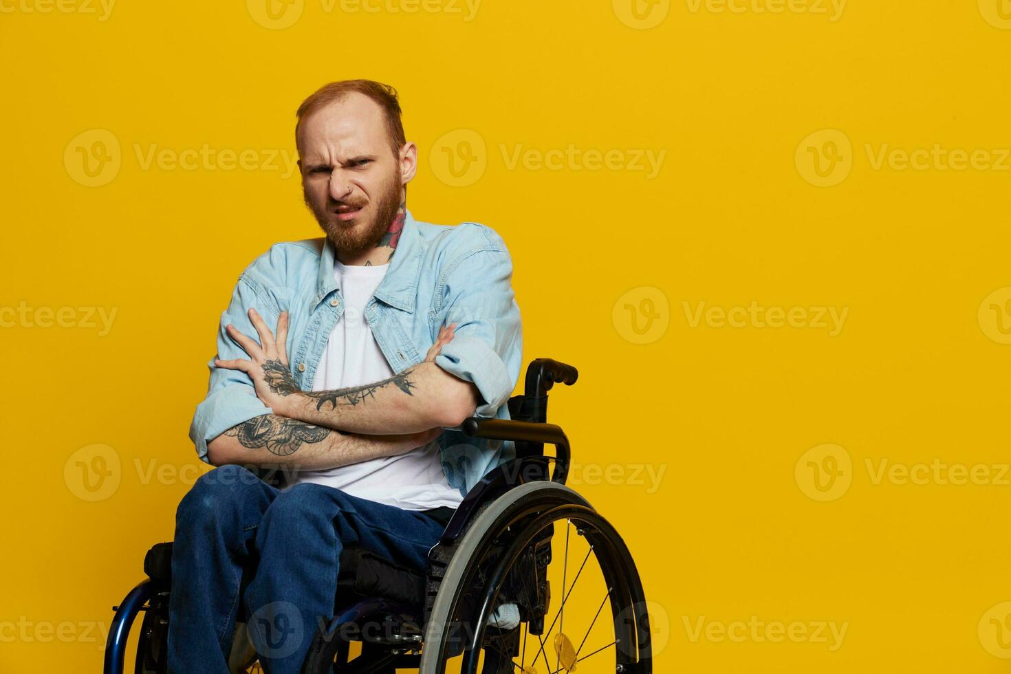 A man in a wheelchair problems with the musculoskeletal system looks at the camera is not satisfied, with tattoos on his arms sits on a yellow studio background, health concept man with disabilities photo