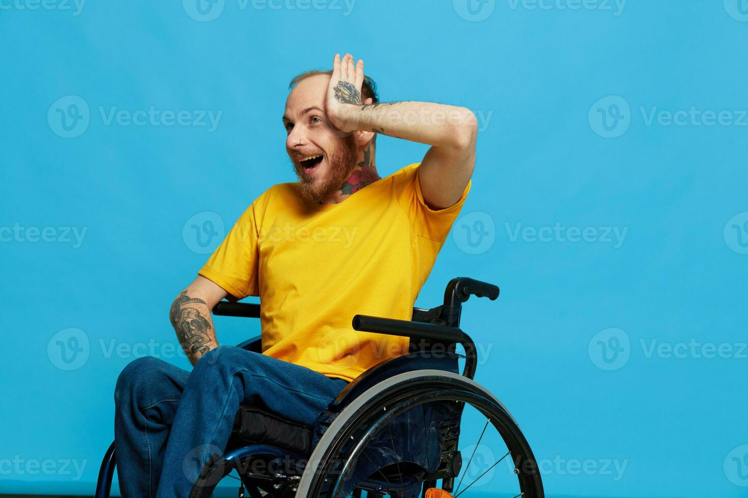 A man in a wheelchair surprised and open mouth in a t-shirt with tattoos on his arms sits on a blue studio background, a full life, a real person photo