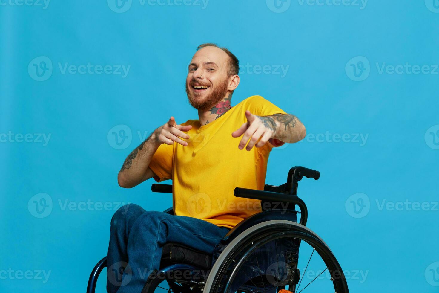 A man in a wheelchair smile looks at the camera in a t-shirt with tattoos on his arms sits on a blue studio background, a full life, a real person photo