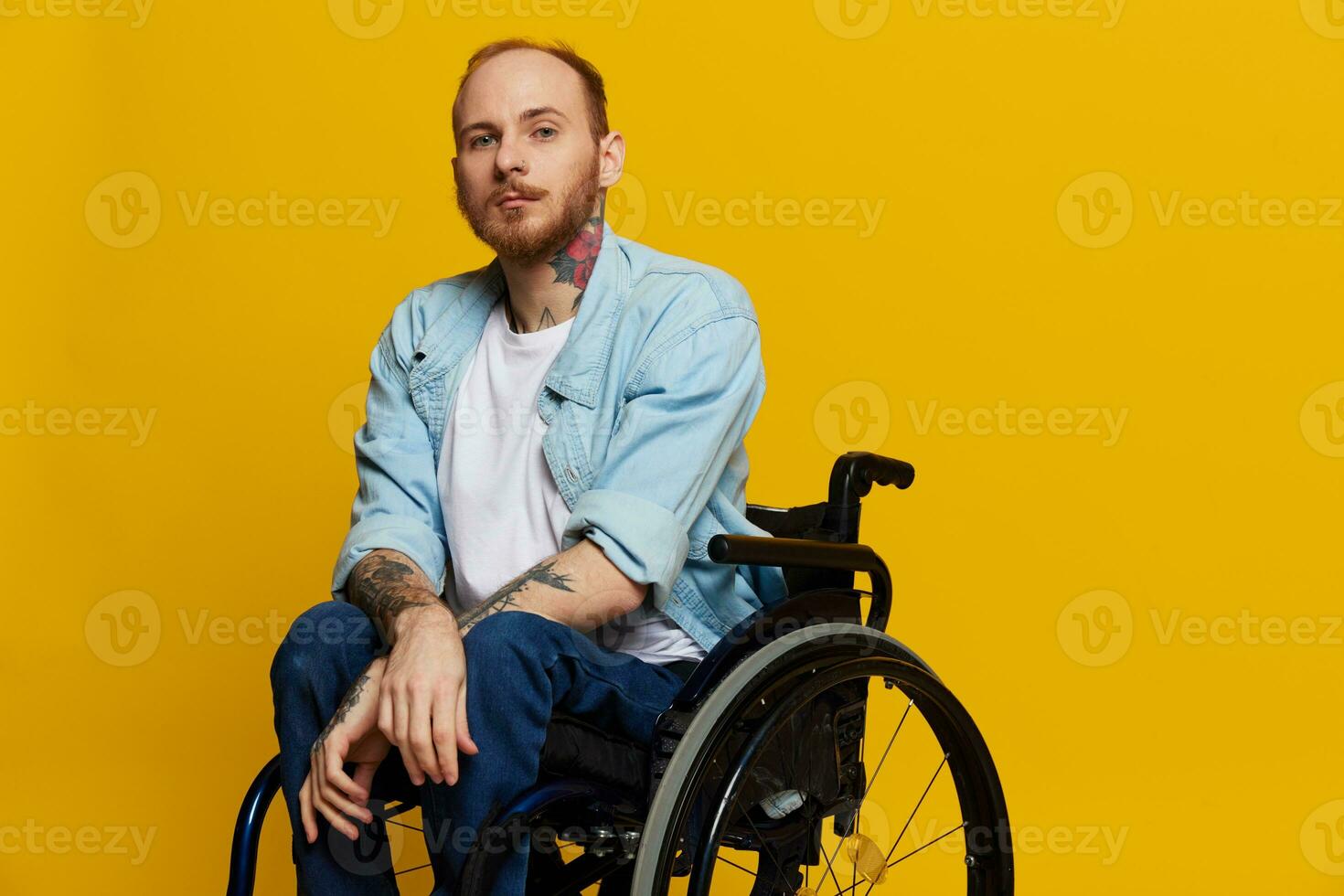 A man in a wheelchair looks at the camera, with tattoos on his arms sits on a yellow studio background, the concept of health is a person with disabilities photo