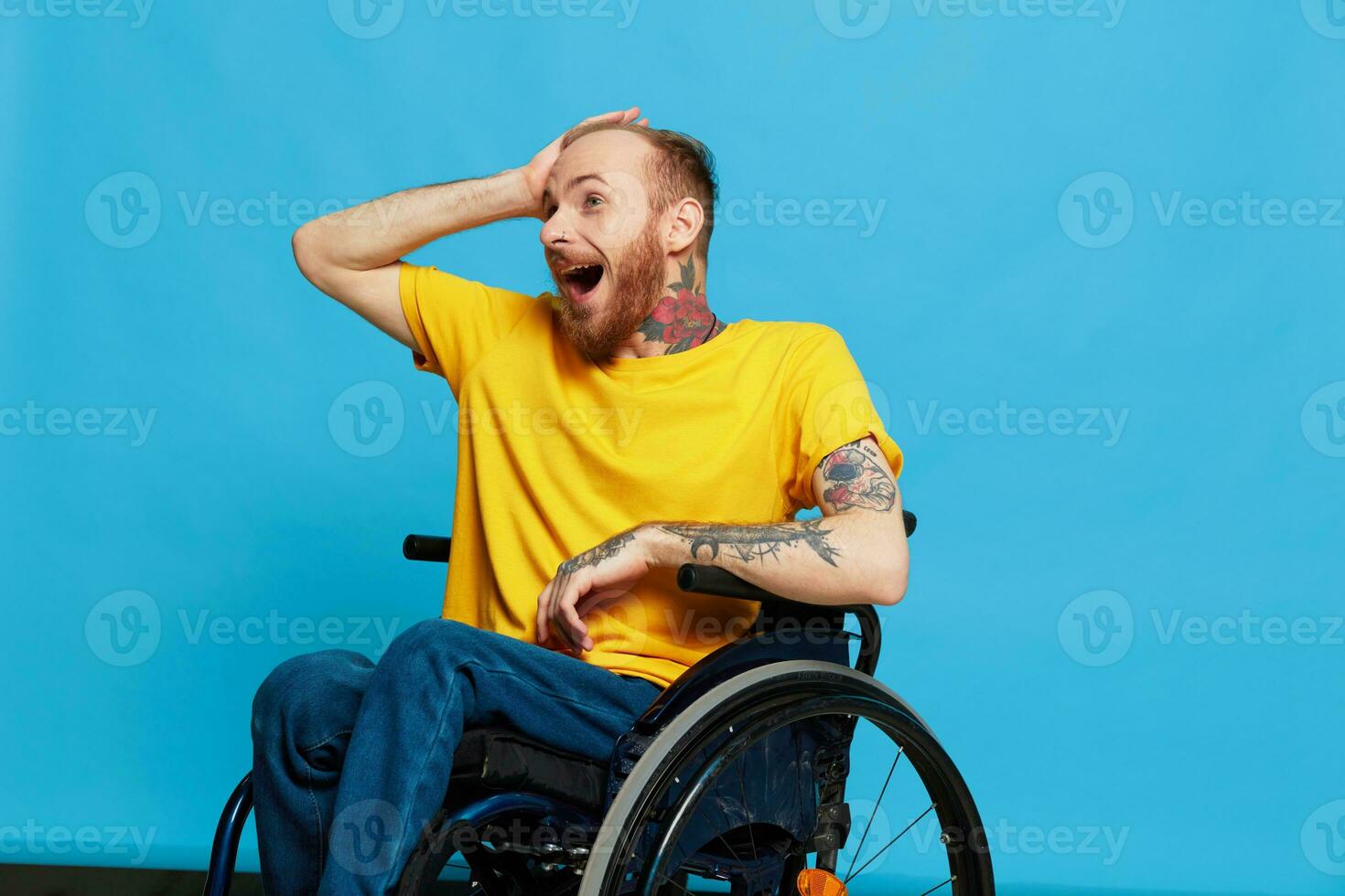 A man in a wheelchair surprised and open mouth in a t-shirt with tattoos on his arms sits on a blue studio background, a full life, a real person photo
