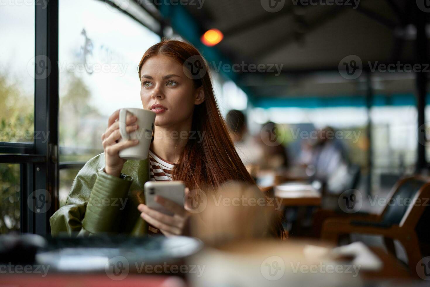 Woman sitting in a cafe drinking coffee from a mug with phone in hand, blogger photographing food content for social media, portrait of a stylish girl with red hair in the autumn photo