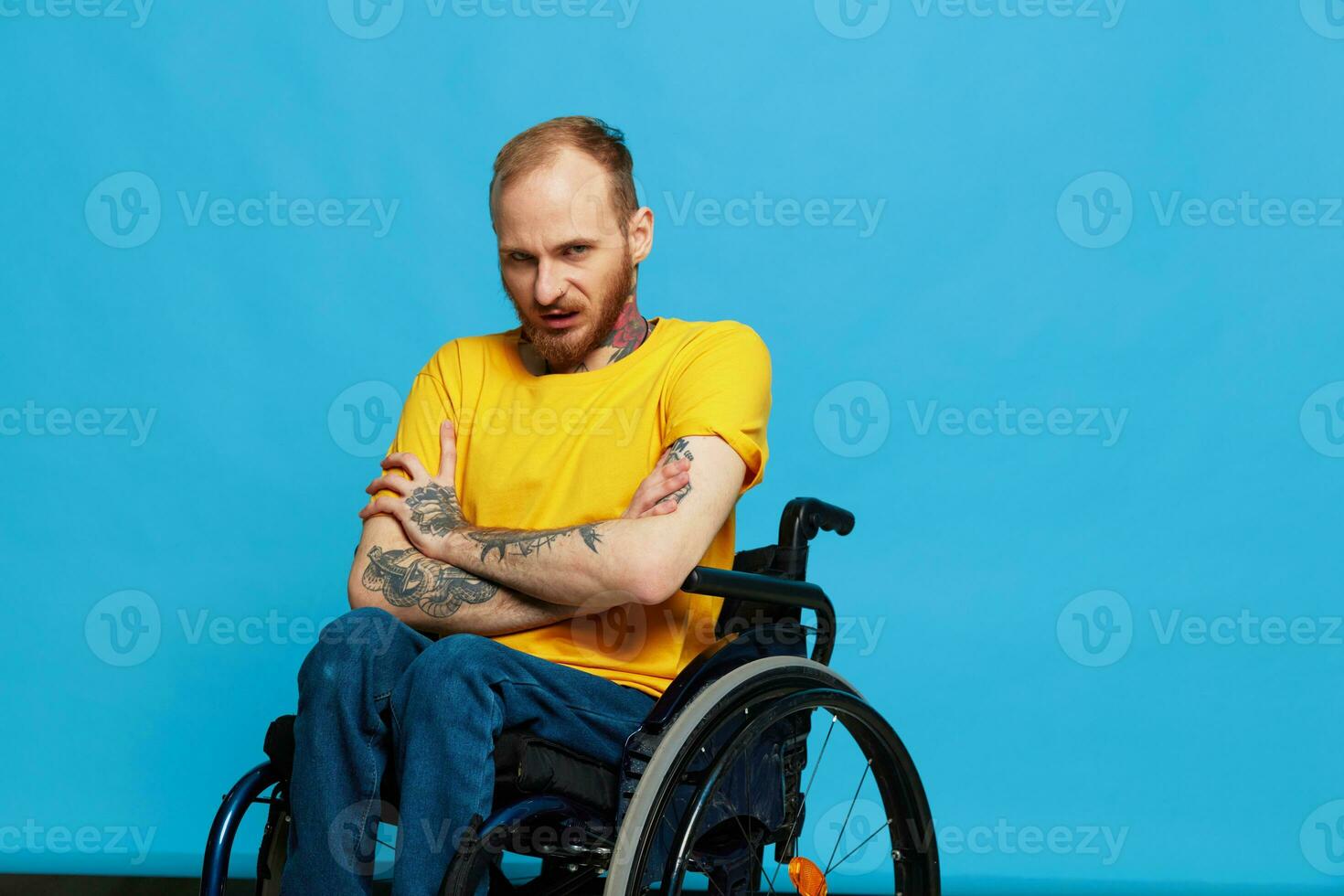 A man in a wheelchair in a T-shirt with tattoos on his arms sits on a blue studio background, a full life, a real person photo