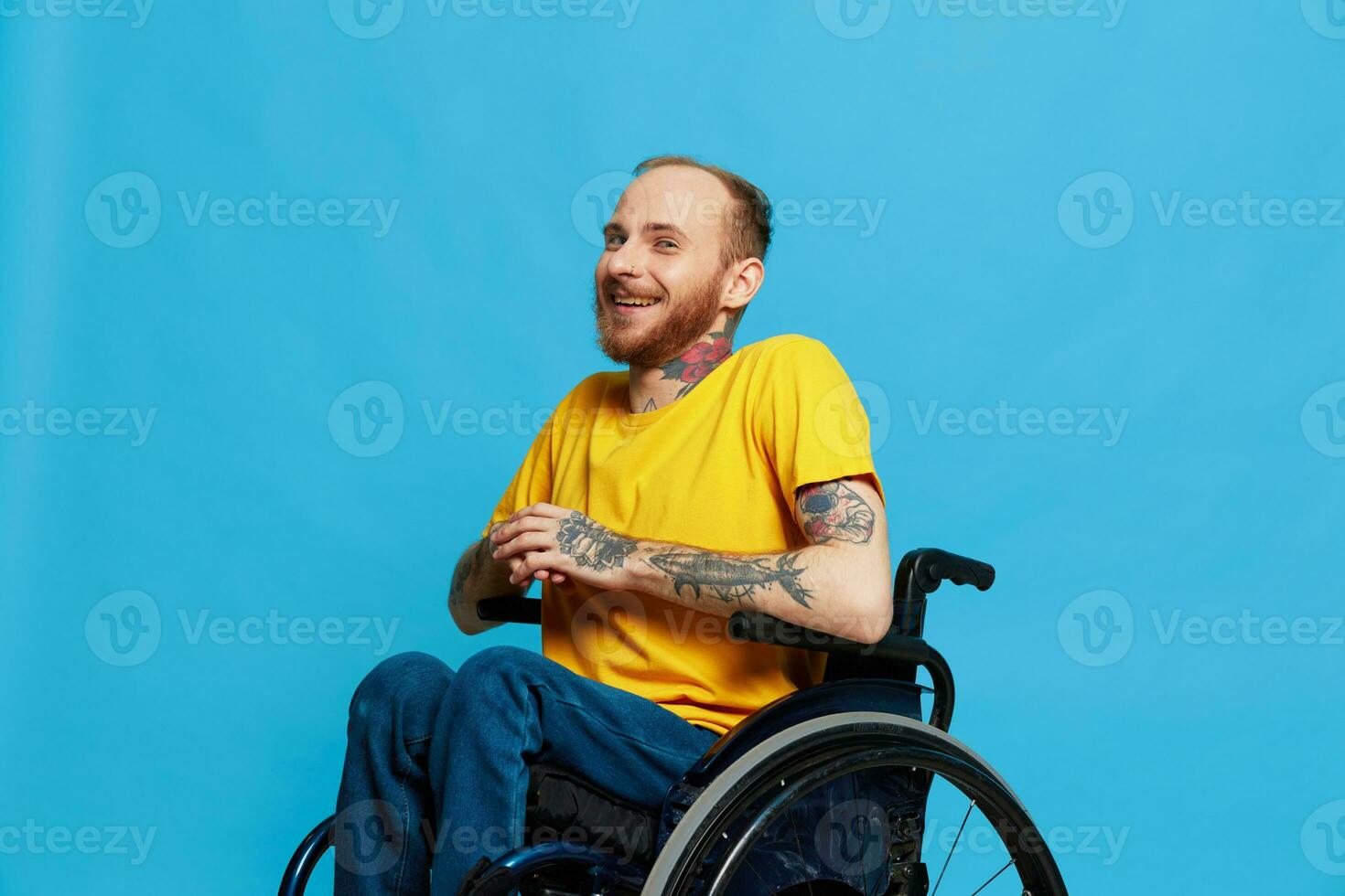 A man in a wheelchair smile looks at the camera in a t-shirt with tattoos on his arms sits on a blue studio background, a full life, a real person photo