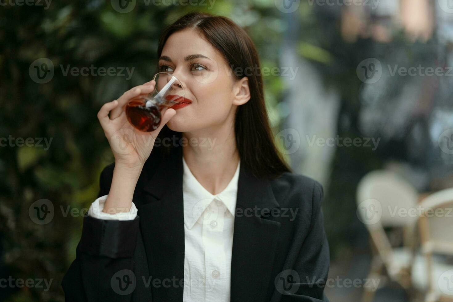 Business brunette woman with red lips smile with teeth drinks tea in a cafe from a Turkish glass mug in a white shirt and black fashion jacket on the street, summer travel, vacation in the city photo