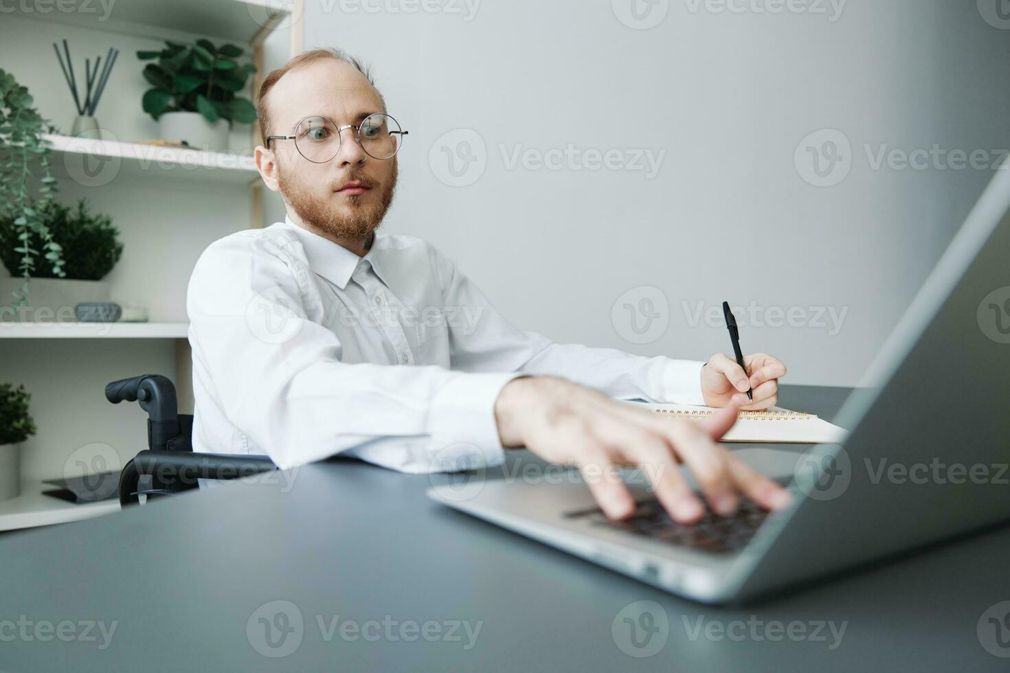 A man wheelchair businessman in the office works at a laptop, writing down ideas and a plan in a notebook, thoughtfulness, integration into society, the concept of working a person with disabilities photo