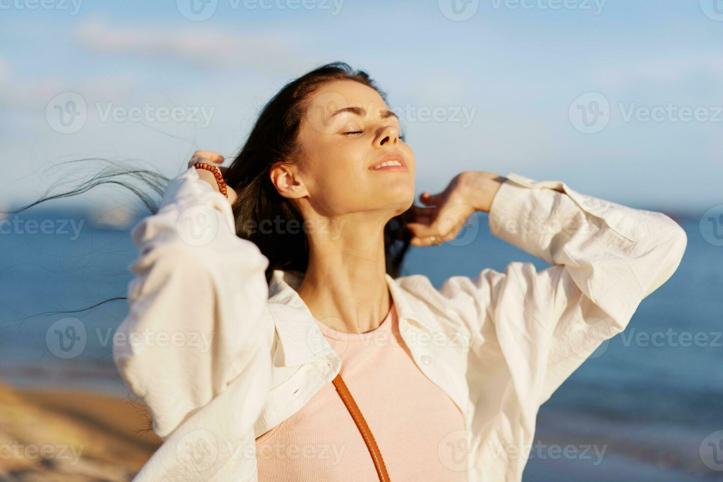 A girl with her eyes closed in the sun against the ocean smile with teeth, flying hair, tanned skin, rest, the concept of skin care in summer and spring. photo