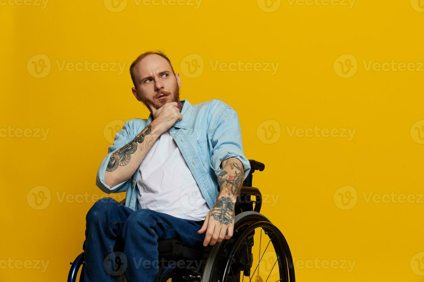A man in a wheelchair problems with the musculoskeletal system looks at the camera thoughtfully with tattoos on his arms sits on a yellow studio background, health concept a person with disabilities photo