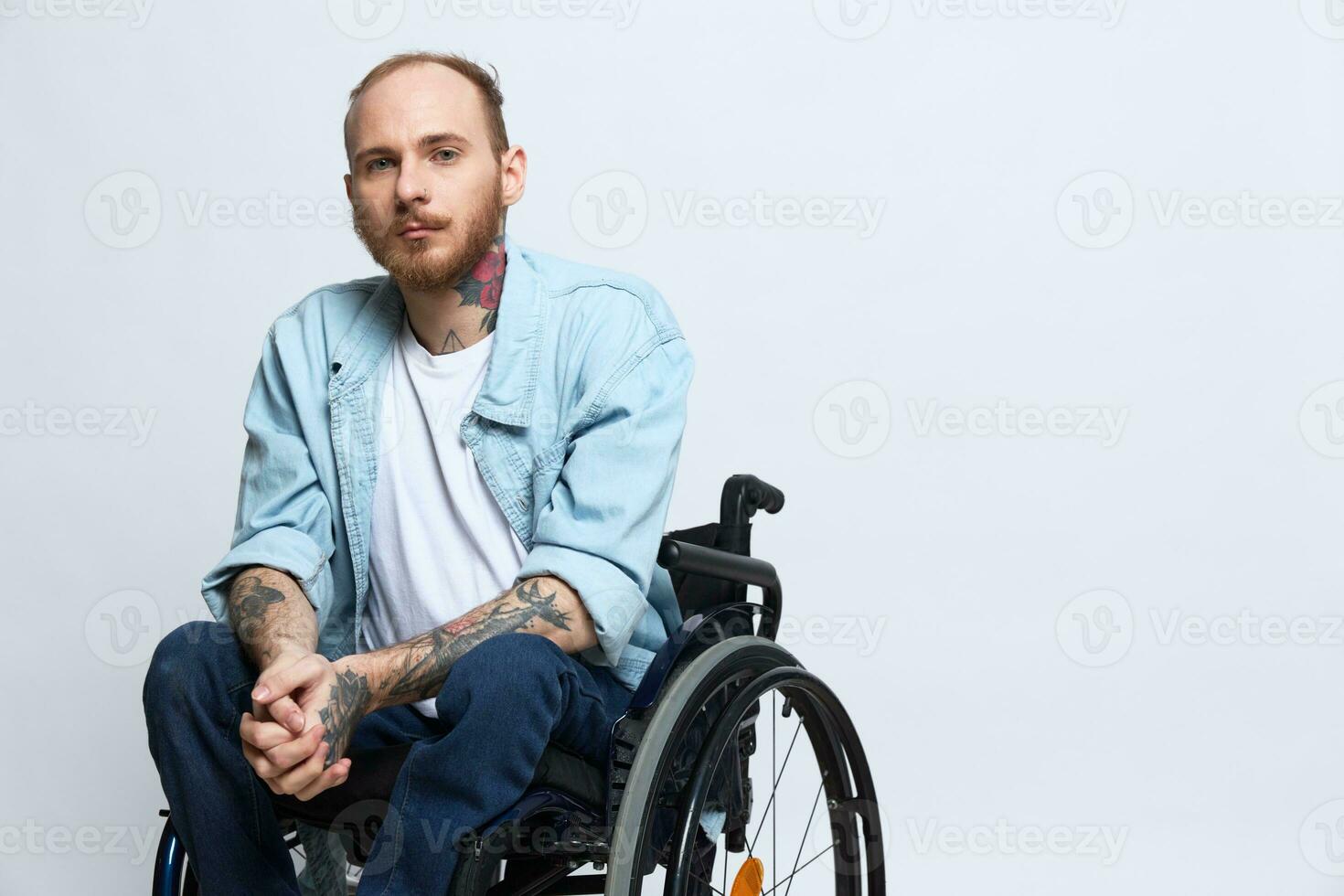 A man in a wheelchair looks thoughtfully at the camera, copy space, with tattoos on his arms sits on a gray studio background, the concept of health a person with disabilities, a real person photo