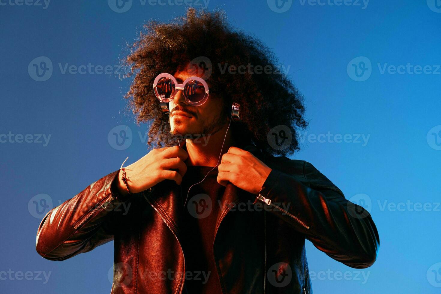 Portrait of a stylish man with curly hair with glasses and headphones on a blue background multinational, colored light, black leather jacket trend, modern concept. photo