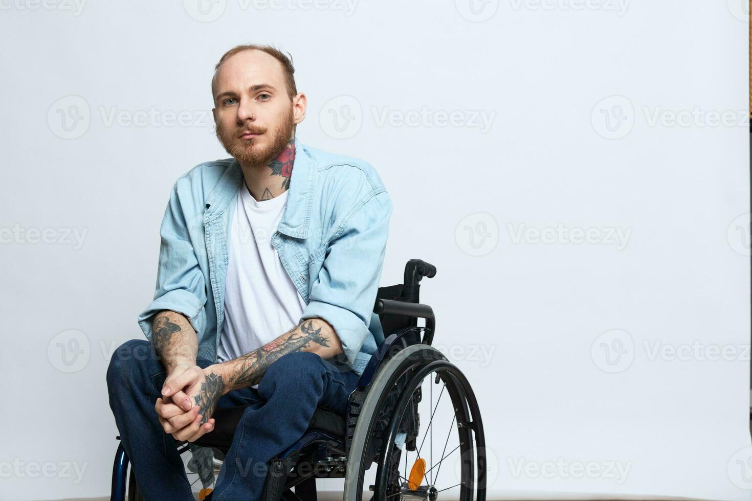 A man in a wheelchair looks thoughtfully at the camera, copy space, with tattoos on his arms sits on a gray studio background, the concept of health a person with disabilities, a real person photo