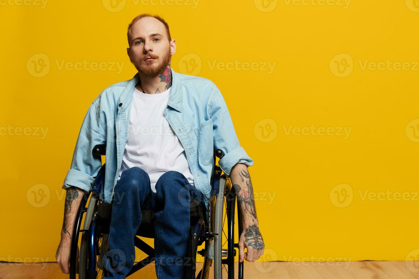 A man in a wheelchair looks at the camera, with tattoos on his arms sits on a yellow studio background, the concept of health is a person with disabilities photo