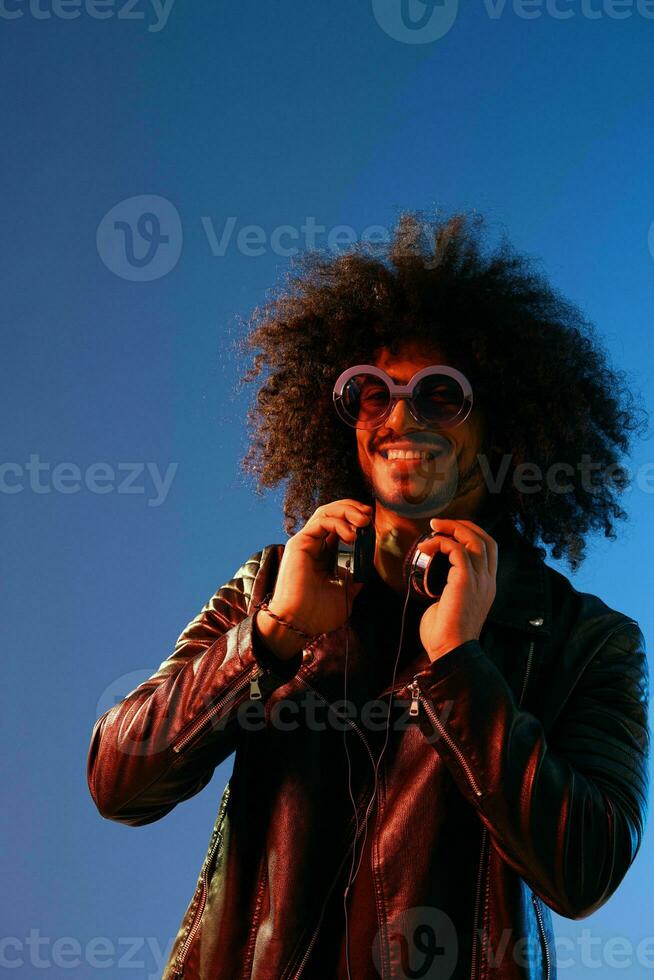 Portrait of a stylish man with curly hair with glasses and headphones on a blue background multinational, colored light, black leather jacket trend, modern concept. photo