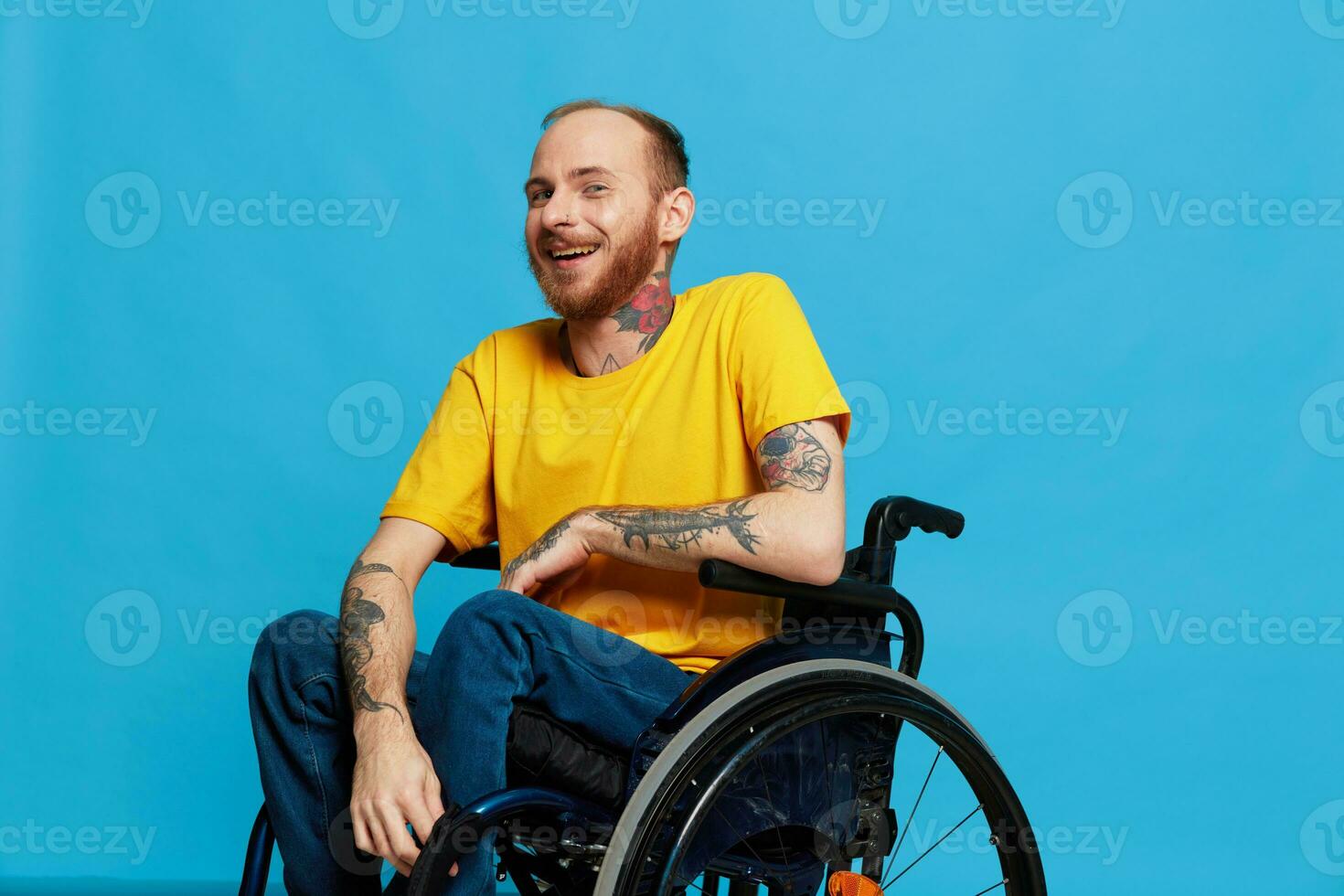 A man in a wheelchair smile looks at the camera in a t-shirt with tattoos on his arms sits on a blue studio background, a full life, a real person photo