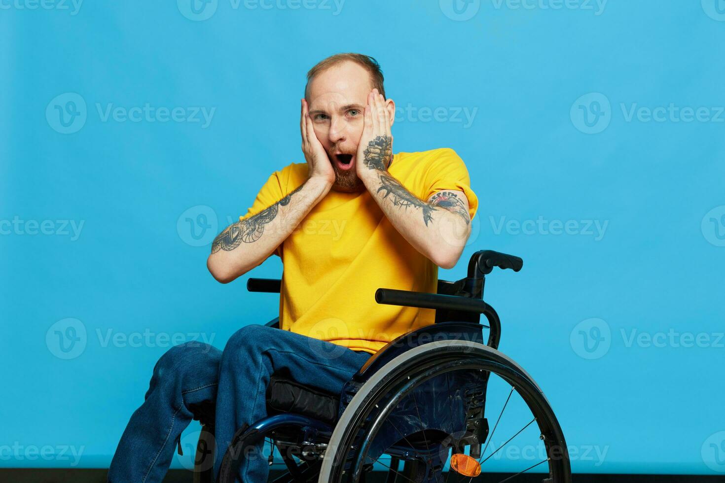 A man in a wheelchair surprised and open mouth in a t-shirt with tattoos on his arms sits on a blue studio background, a full life, a real person photo