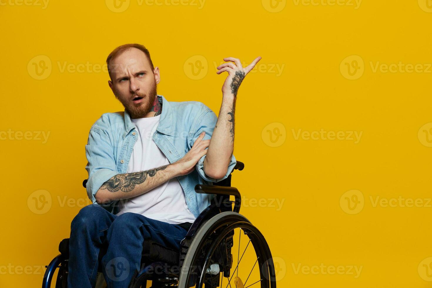 A man in a wheelchair problems with the musculoskeletal system looks at the camera shows a finger on, with tattoos on his hands sits on a yellow studio background, health concept man with disabilities photo