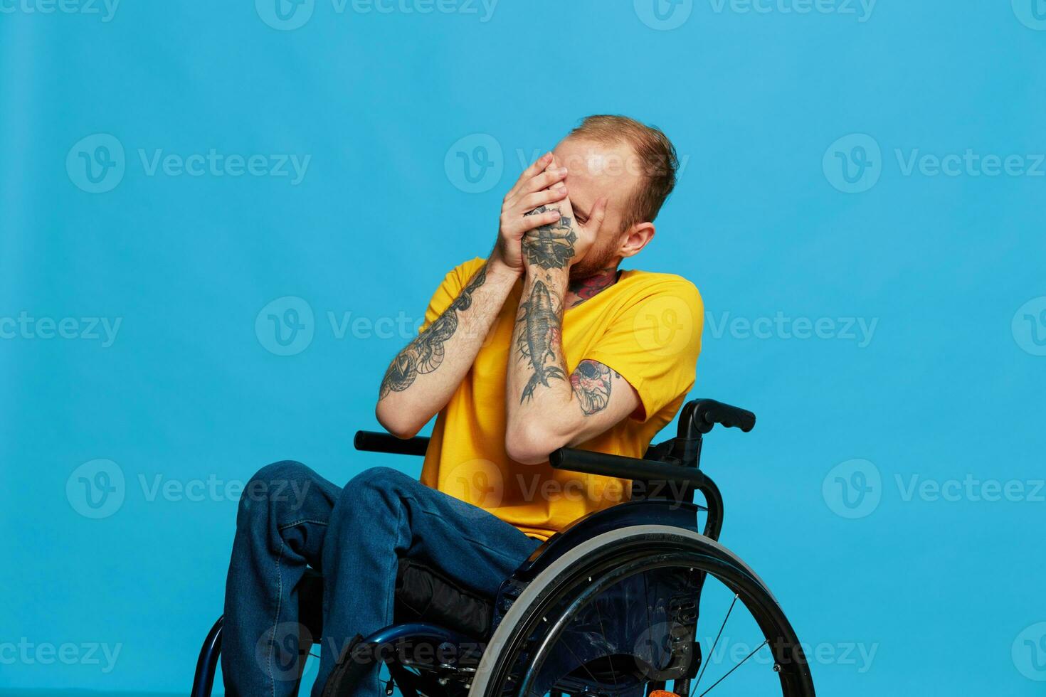 A man in a wheelchair smile looks at the camera in a t-shirt with tattoos on his arms sits on a blue studio background, a full life, a real person photo