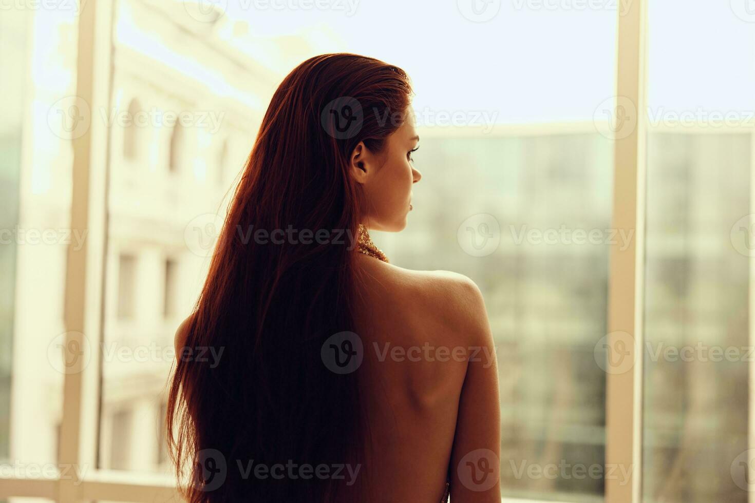 Woman in gold sequined dress standing by the window posing and looking at the city in a stylish look with long hair, designer dress with open back, party look photo