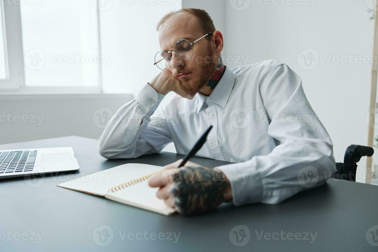 A man in a wheelchair, a businessman in the office works at a laptop, writing down a plan in a notebook, thoughtfulness, integration into society, the concept of working a person with disabilities photo
