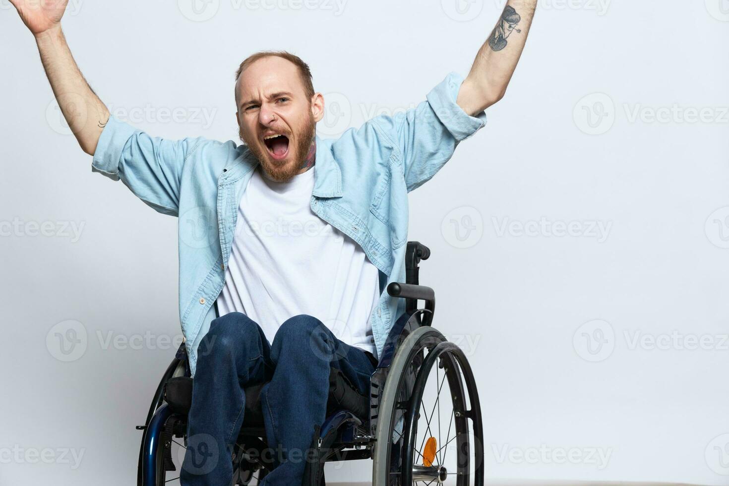 A man in a wheelchair looks at the camera anger and aggression, with tattoos on his arms sits on a gray studio background, health concept man with disabilities photo