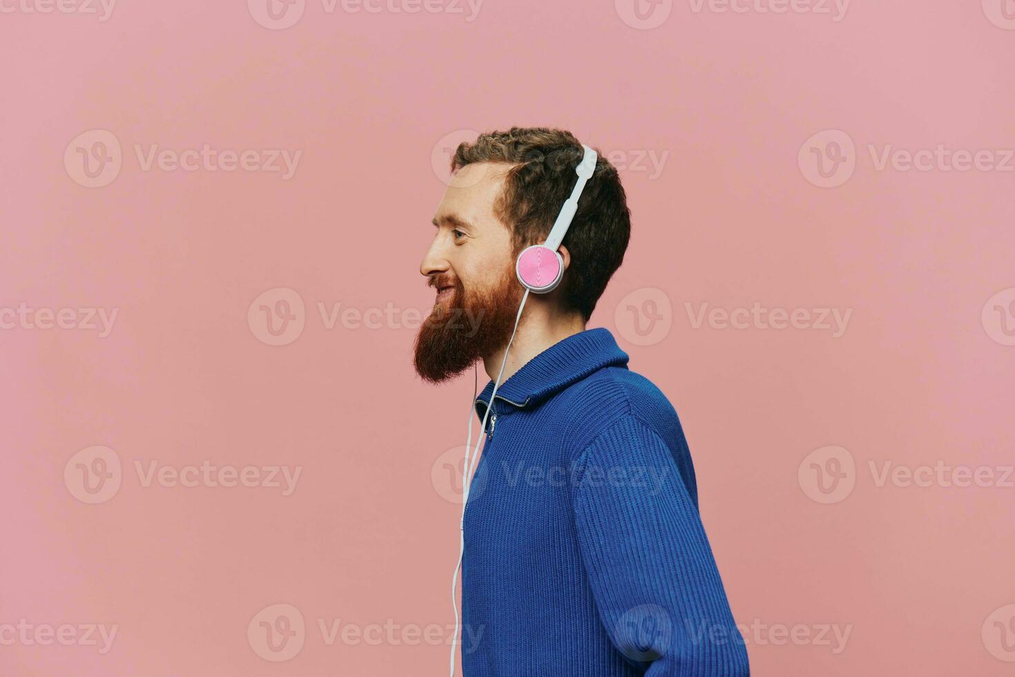 Portrait of a redheaded man wearing headphones smiling and dancing, listening to music on a pink background. A hipster with a beard. photo