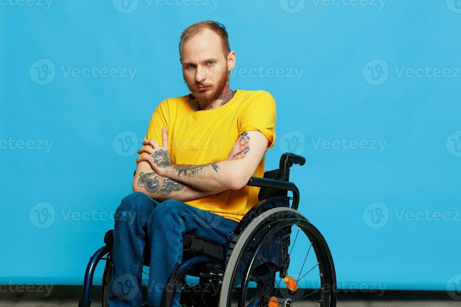 A man in a wheelchair in a T-shirt with tattoos on his arms sits on a blue studio background, a full life, a real person photo