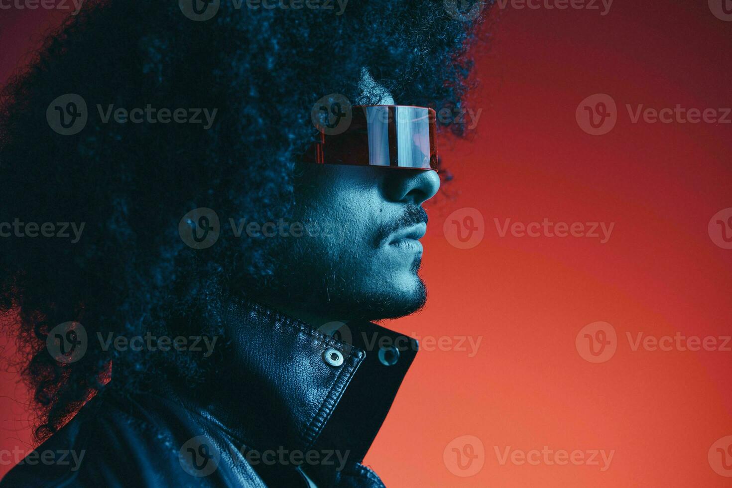 Portrait of fashion man with curly hair on red background with stylish glasses, multicultural, colored light, black leather jacket trend, modern concept. photo