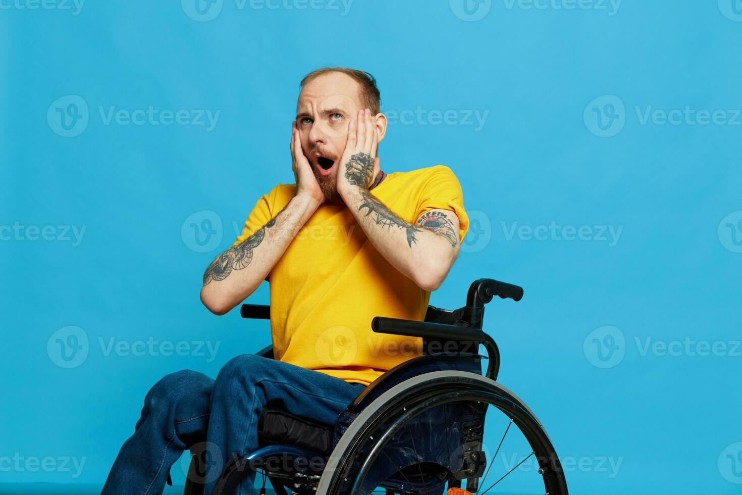 A man in a wheelchair surprised and open mouth in a t-shirt with tattoos on his arms sits on a blue studio background, a full life, a real person photo