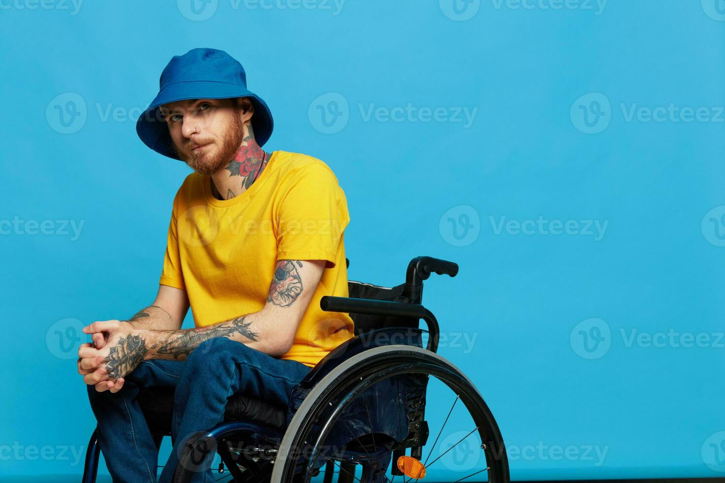 A man in a wheelchair problems with the musculoskeletal system looks at the camera in a t-shirt with tattoos on his arms sits on a blue studio background, full life, real person, health concept photo