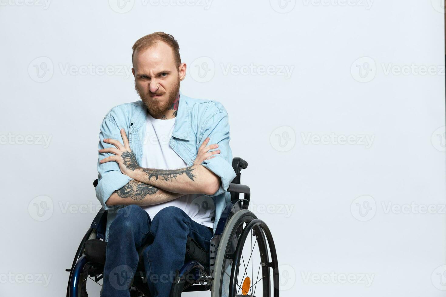 A man in a wheelchair looks at the camera anger and aggression, with tattoos on his arms sits on a gray studio background, health concept man with disabilities photo
