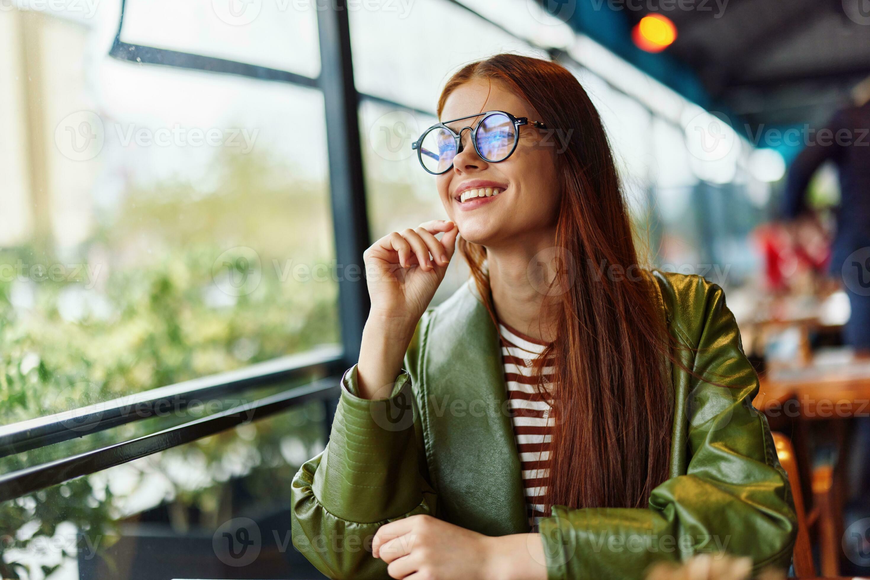 Beautiful woman with red hair and glasses sits in the city in a cafe and  looks out the window smile with teeth, female freelance blogger 25716503  Stock Photo at Vecteezy