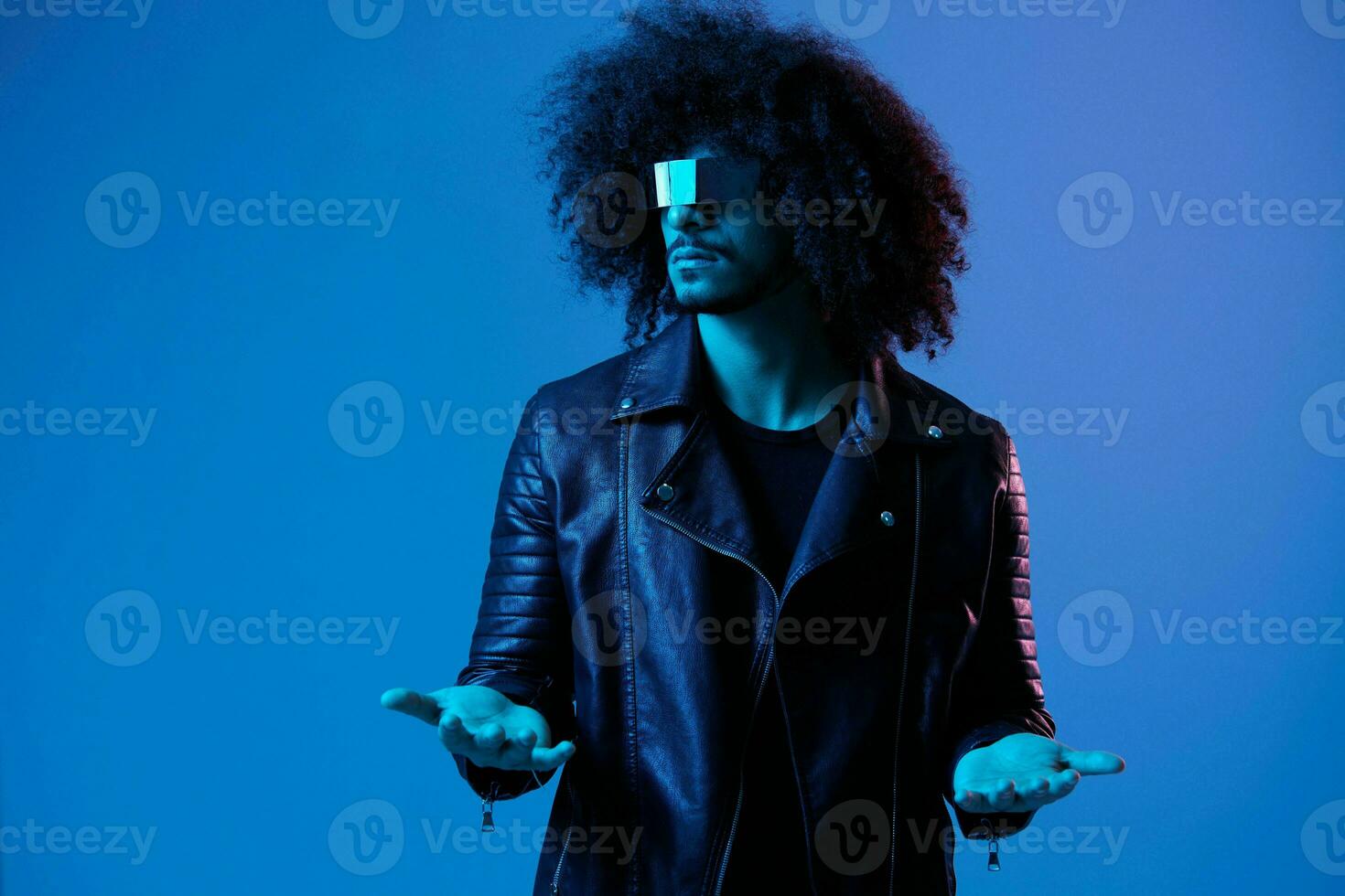 Portrait of fashion man with curly hair with stylish glasses on blue background multinational, colored light, black leather jacket trend, modern concept. photo