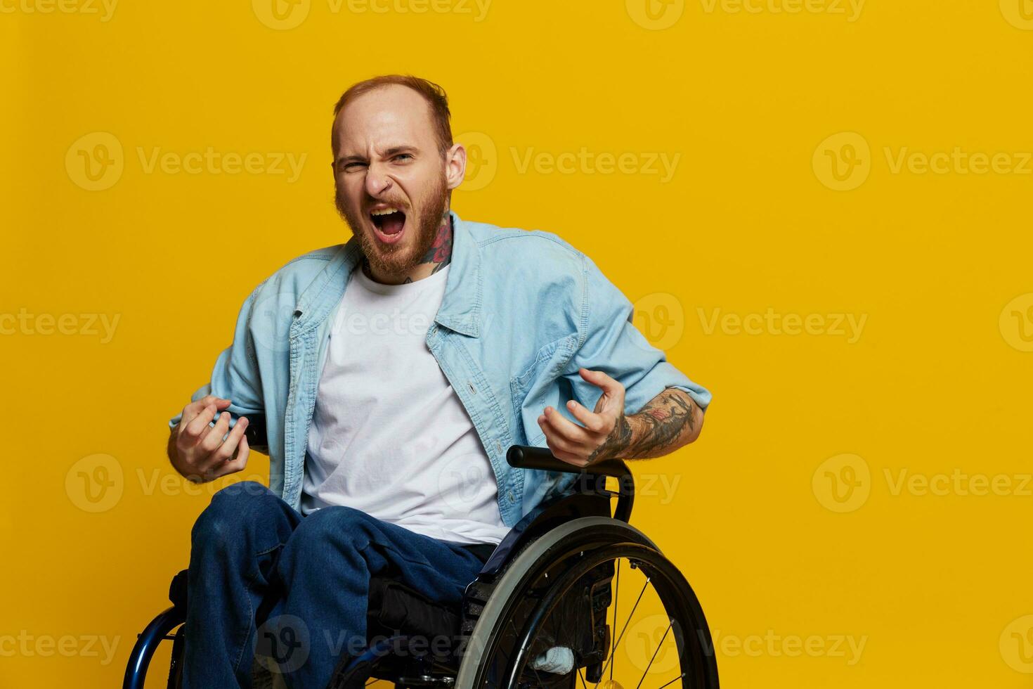 man in a wheelchair screams angrily, shows fist looks at the camera is not pleased, with tattoos on his hands sits on yellow background of the studio, the concept of health is person with disabilities photo
