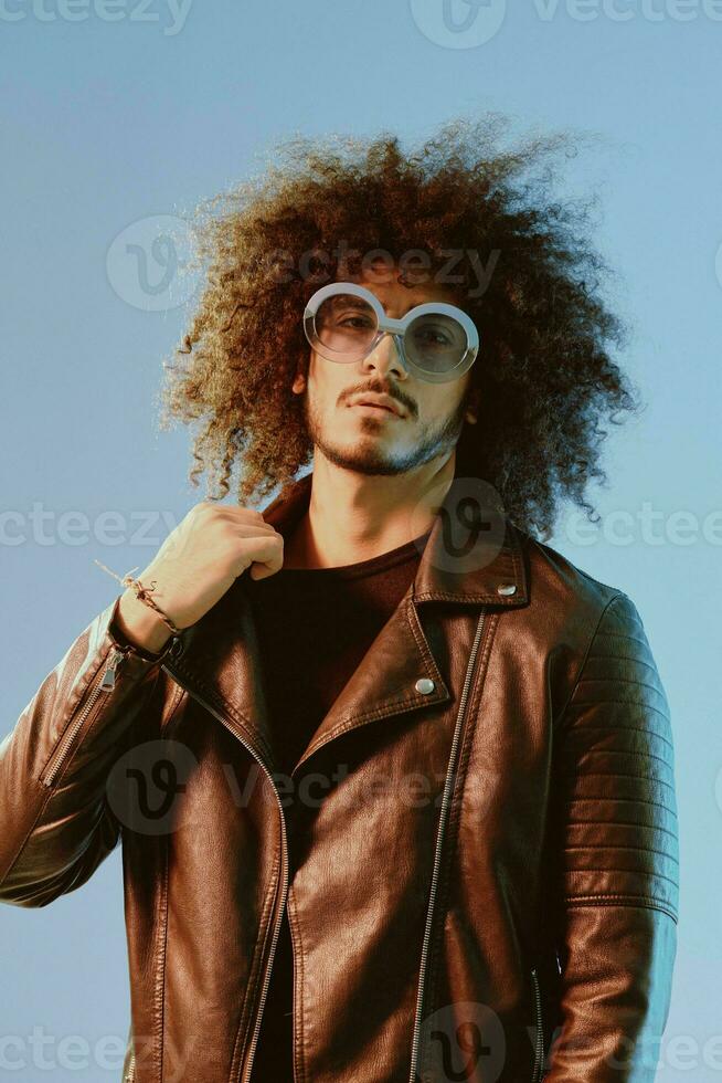 Portrait of a stylish man with curly hair with glasses smile on blue background multinational, color light, black leather jacket trend, modern concept. photo