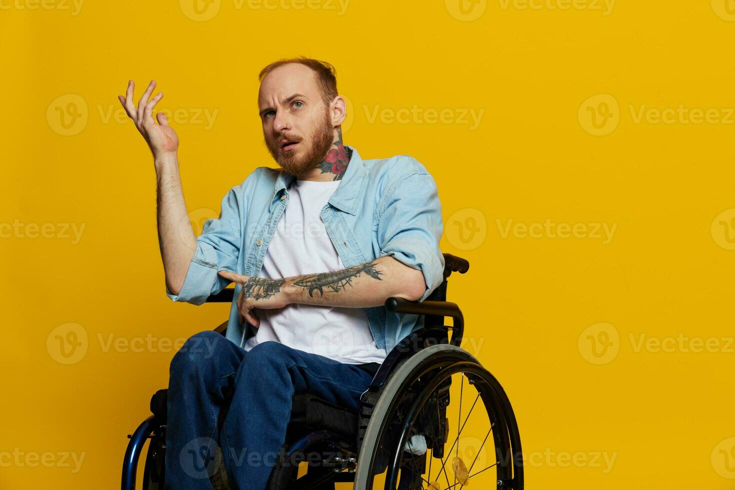 A man in a wheelchair problems with the musculoskeletal system looks at the camera is not satisfied, with tattoos on his arms sits on a yellow studio background, health concept man with disabilities photo