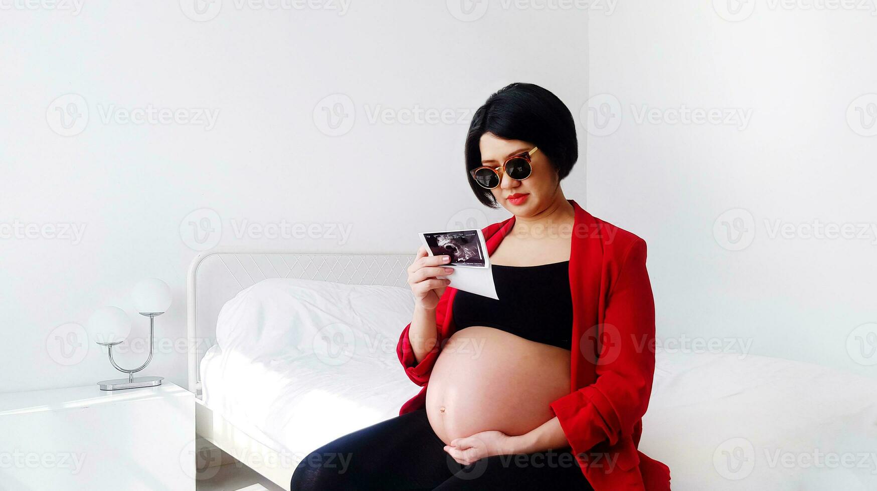 Beautiful and pretty pregnant woman wearing black glasses, sitting on bed, looking at her baby's film picture and touching her belly. Pregnancy 7-8 months, motherhood, love, expectation and care. photo