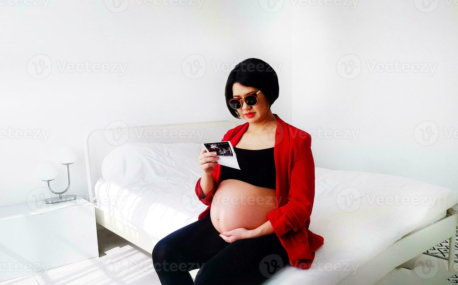 Beautiful and pretty pregnant woman wearing black glasses, sitting on bed, looking at her baby's film picture and touching her belly. Pregnancy 7-8 months, motherhood, love, expectation and care. photo