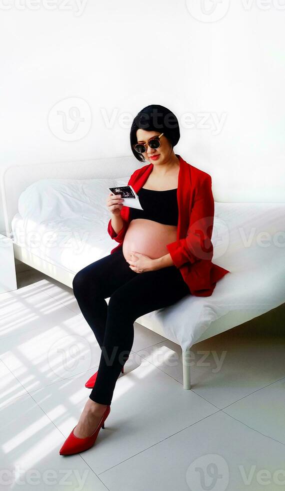 Beautiful or pretty pregnant woman wearing black glasses, sitting on bed, looking at her baby's film picture and touching her belly. Pregnancy 6-8 months, motherhood, love, expectation, care, fashion. photo