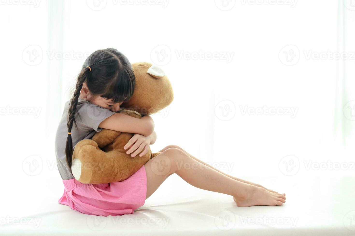 linda pequeño asiático niña con coletas ella se sentó abrazando un marrón osito de peluche oso en el cama. blanco antecedentes. positivo persona.copiar espacio foto