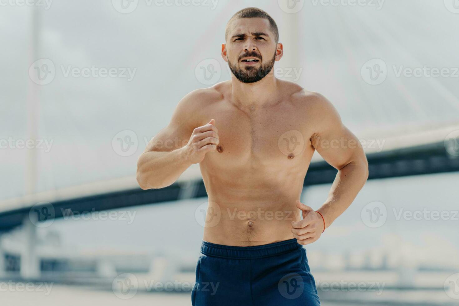 Serious male jogger runs actively, focused on the distance, posing with a naked torso against a blurred background with a bridge. Fit, muscular man trains actively. photo