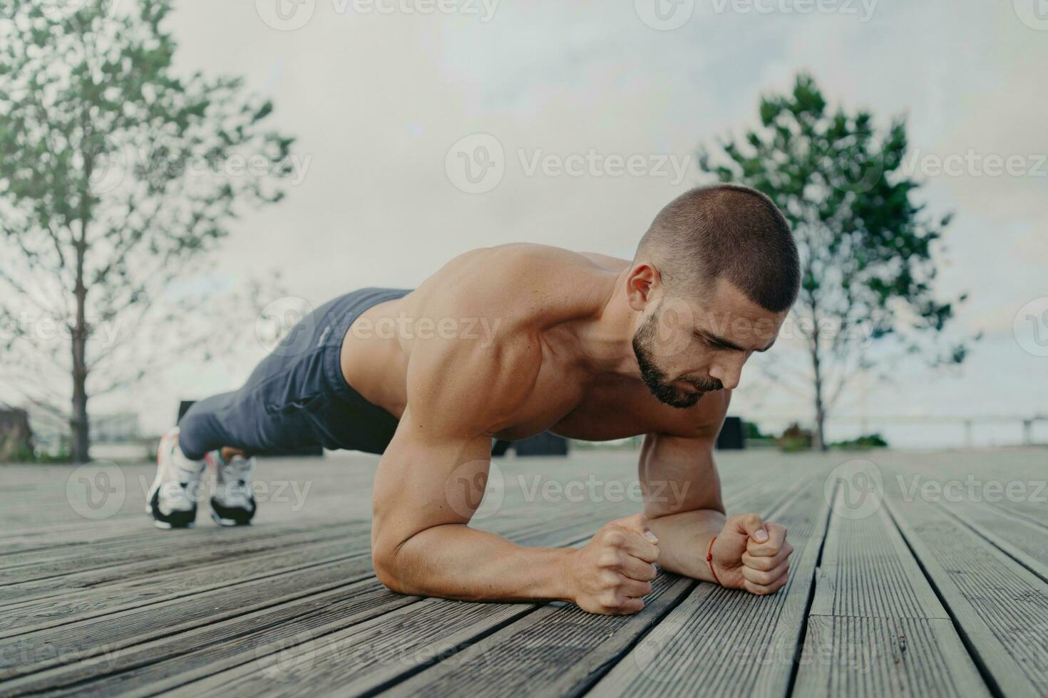 Strong athlete maintains plank pose, prioritizing fitness and health. Concentrated man exercises outdoors, emphasizing abdominal strength. Bodybuilder focuses on core workout. photo