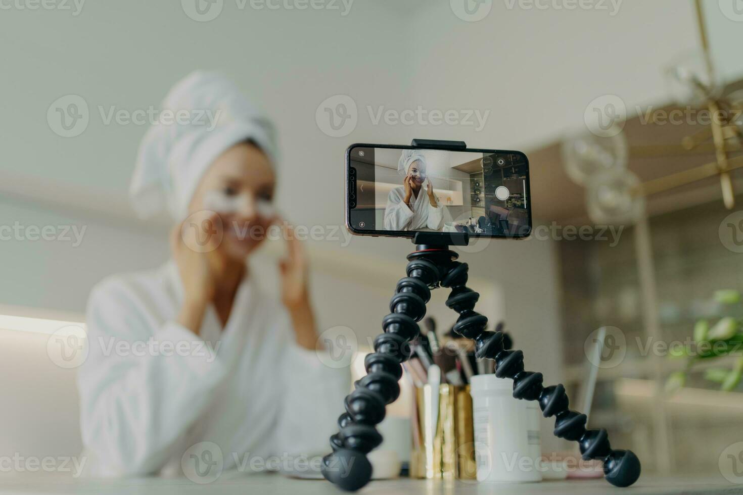 Beauty blogger records under-eye patch tutorial, tackles wrinkles and dark circles, focused on smartphone, bathrobe attire. photo