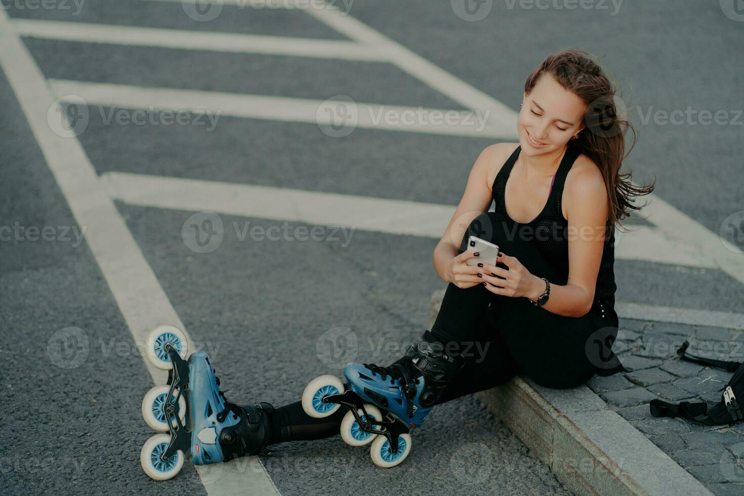 Top view of brunette active young woman uses smartphone for online communication poses on asphalt dressed in black active wear enjoys favorite hobby and free time. Extreme sport and rest concept photo