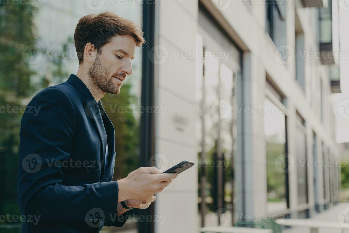 Focused man in dark suit chats, browses on phone near windowed building, typing message photo