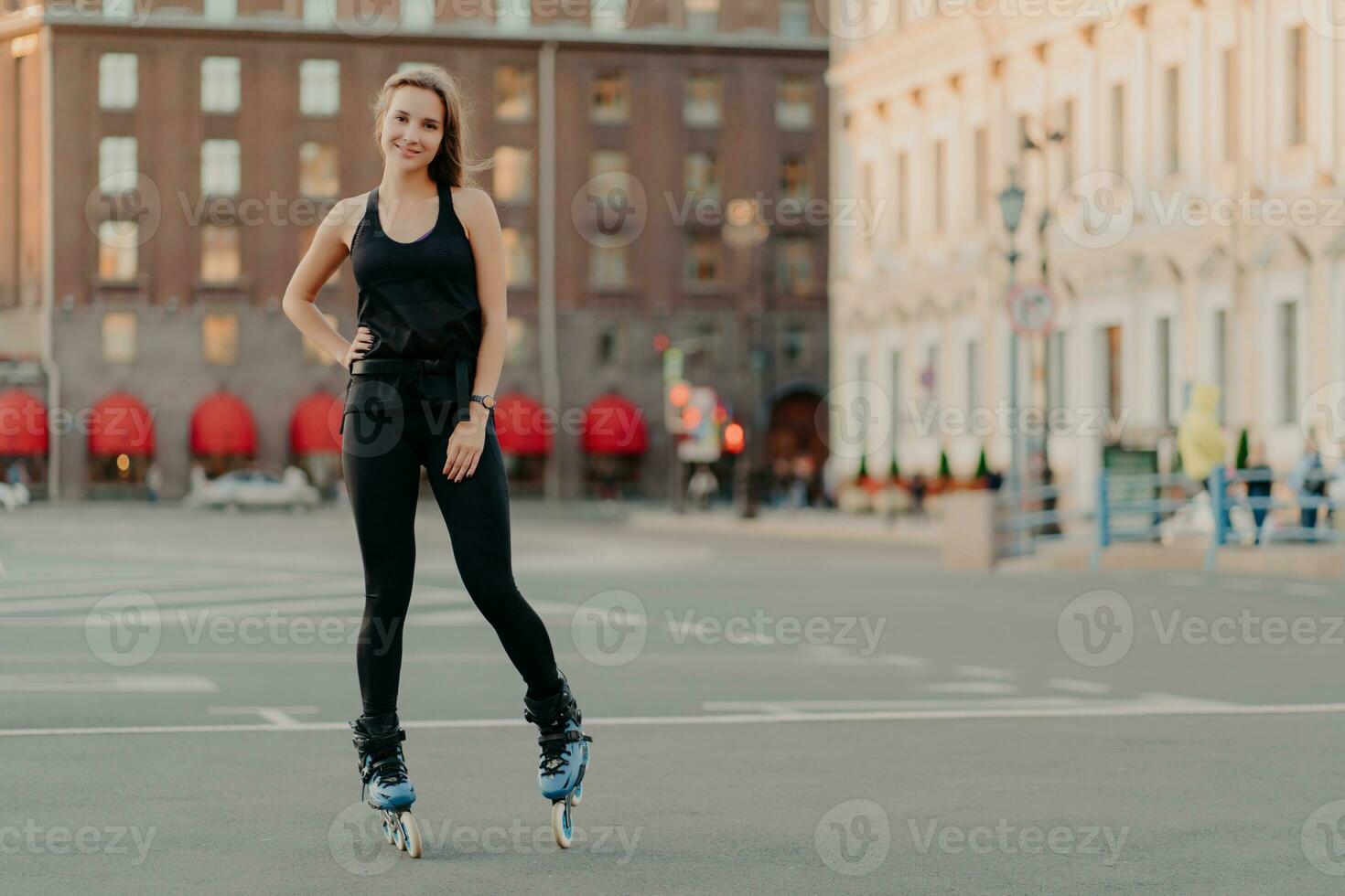 Sport and recreation concept. Horizontal shot of active woman practices rollerblading poses on blades keeps hand on waist blurred city background takes break. Rolleskating after work. Healthy life photo