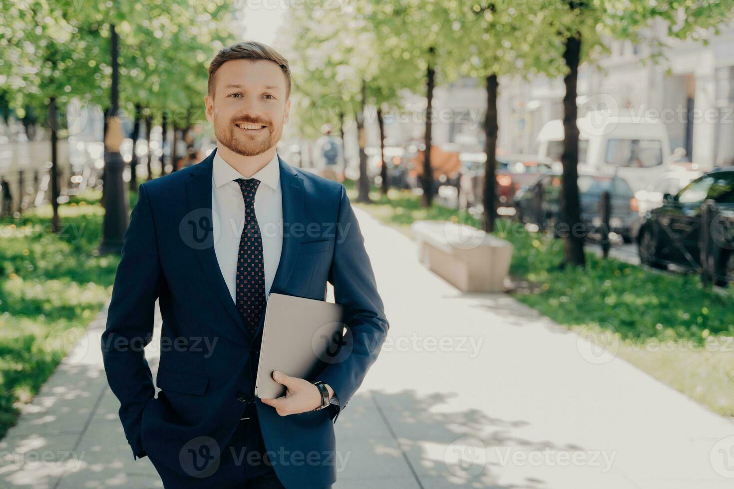 Happy office worker in blue suit holds laptop with data, taking break in park, enjoying fresh air. photo
